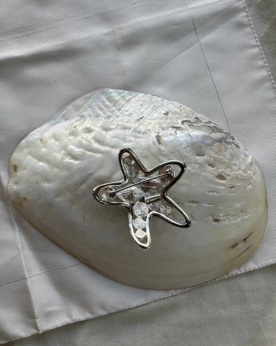 A silver starfish-shaped brooch with clear rhinestones, displayed on top of a white mother-of-pearl clam shell on a white textured fabric background.