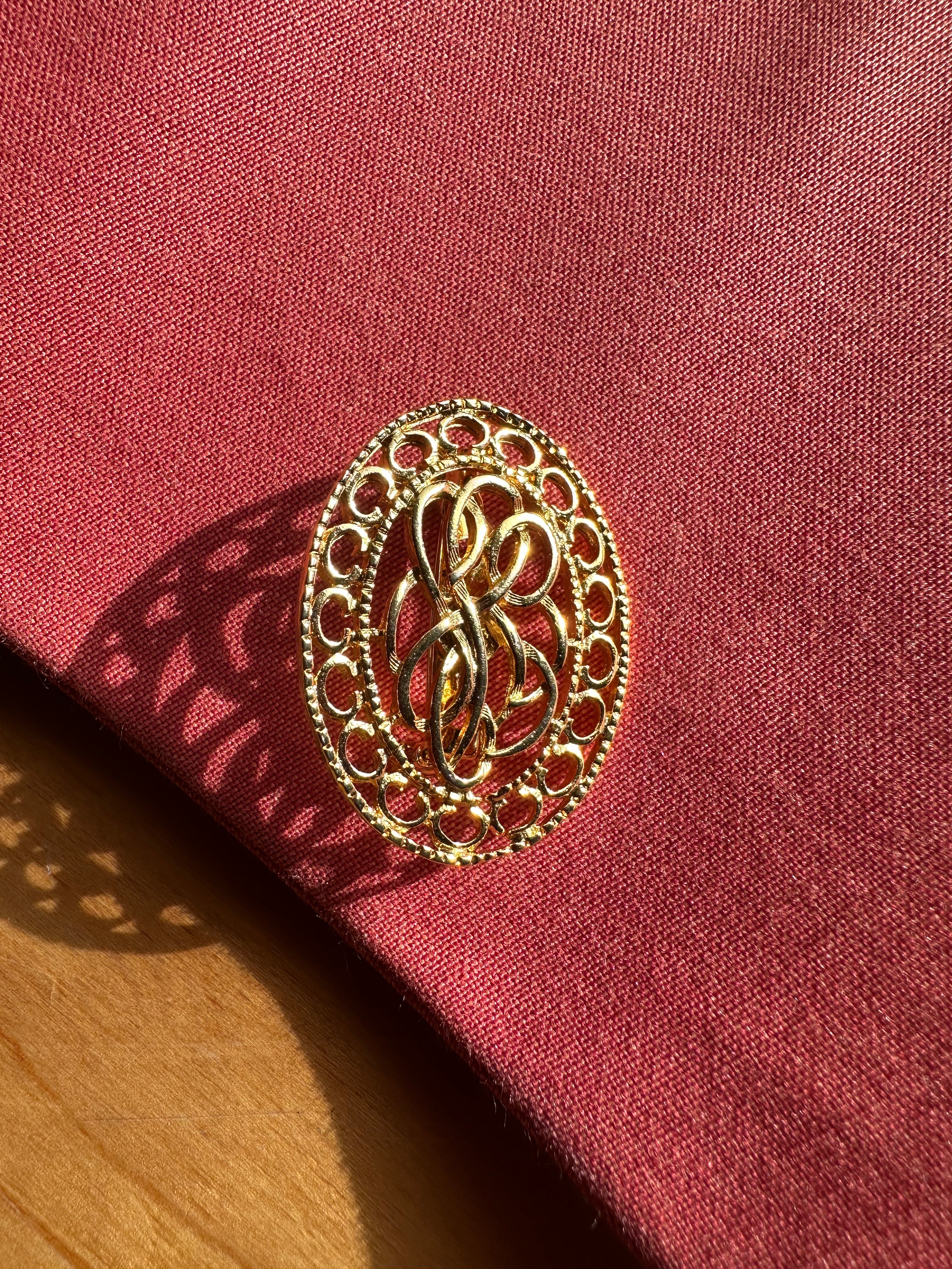 An oval gold filigree brooch with an interlocking monogram design, resting on a textured dark red fabric surface with a wooden surface visible in the lower-left corner.