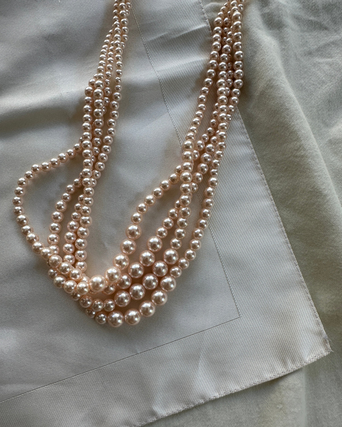 A multi-strand pearl necklace in a champagne or light gold tone, draped elegantly on a textured, light beige fabric background.