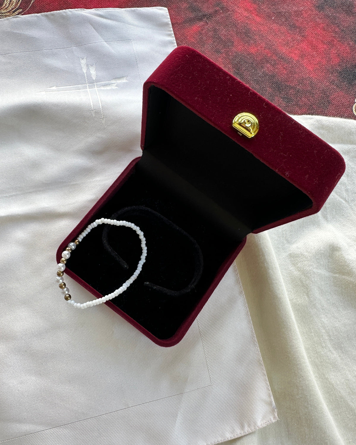 A white beaded bracelet with small gold-toned beads lies inside an open burgundy velvet jewelry box with a black interior and a gold clasp, placed on white fabric.