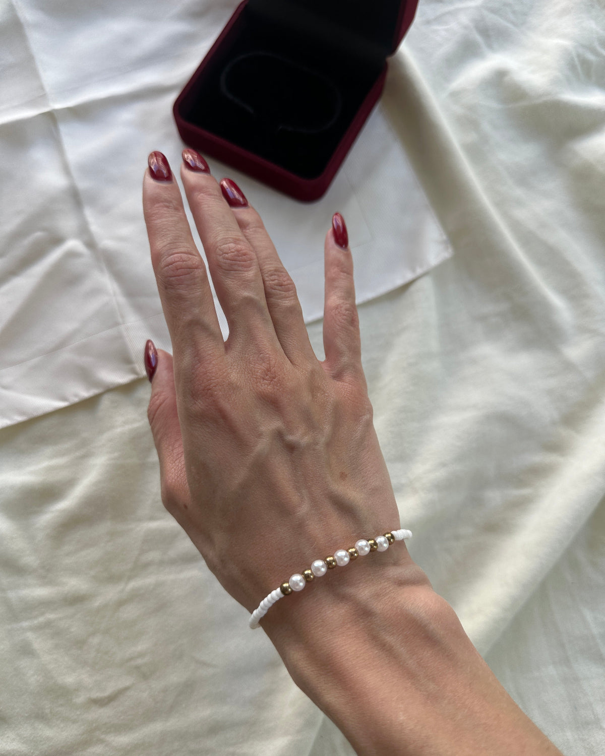 A close-up of a person's hand wearing a delicate white pearl bracelet, with a dark red velvet jewelry box resting on a white fabric background.