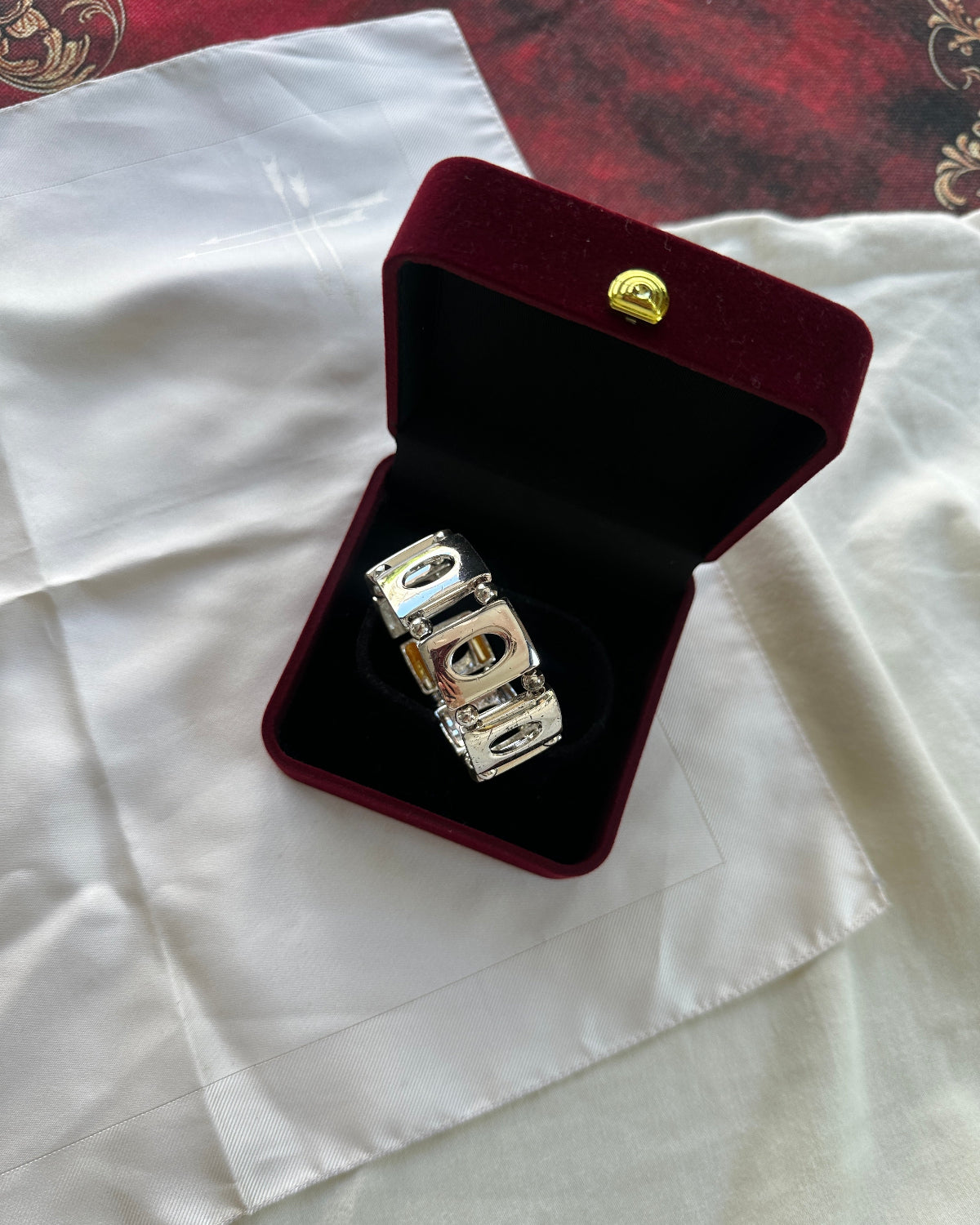 A silver-toned metal bracelet or anklet with a geometric pattern, displayed inside an open dark red velvet jewelry box with a gold clasp, set on a white fabric background.