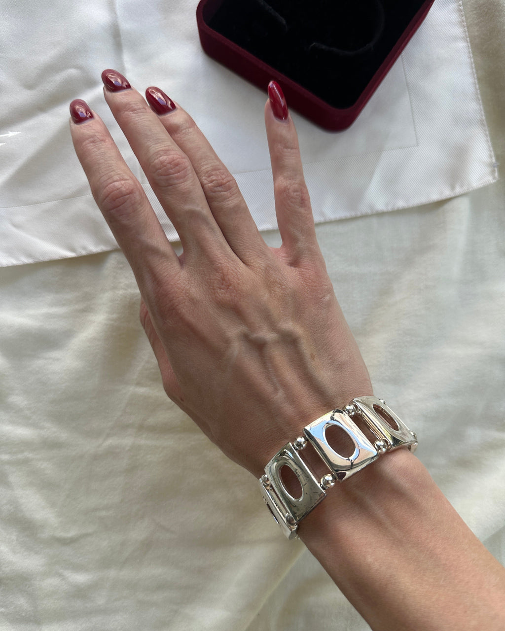 A close-up of a person's hand wearing a chunky silver-tone cuff bracelet with oval cutouts, resting on a white fabric surface next to a dark red jewelry box.