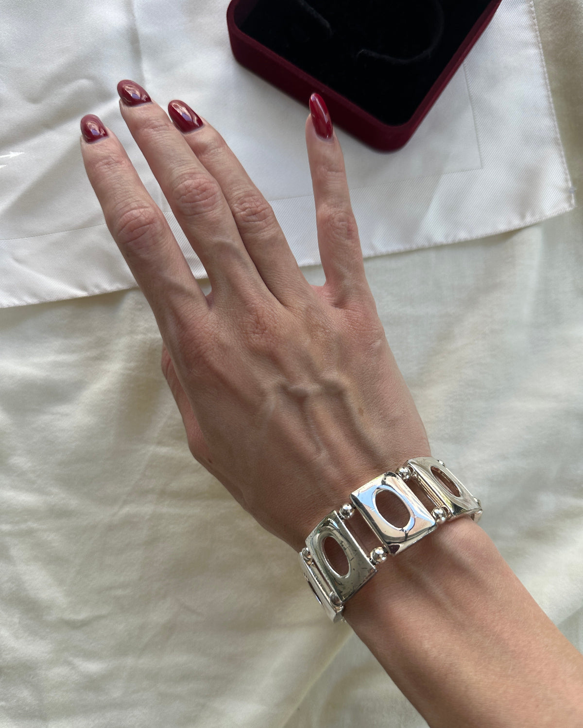 A close-up of a person's hand wearing a chunky silver-tone cuff bracelet with oval cutouts, resting on a white fabric surface next to a dark red jewelry box.