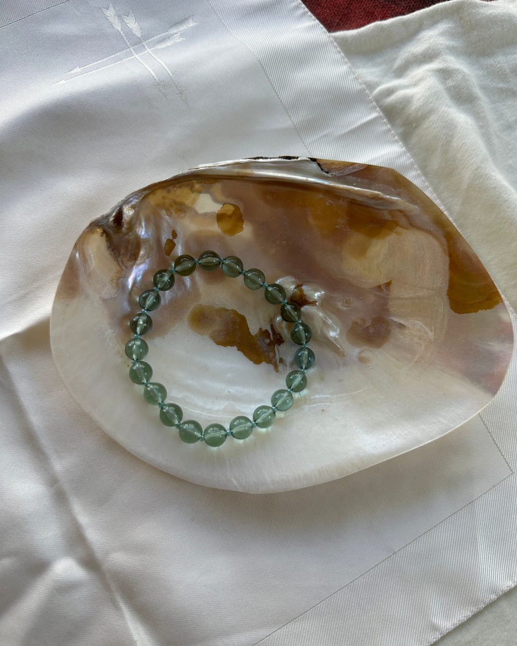 A green beaded bracelet rests inside a polished agate shell dish on a white fabric surface.