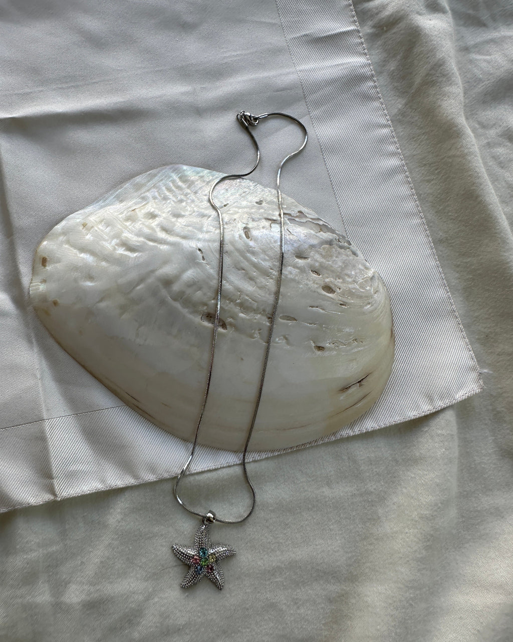 A silver-toned necklace with a starfish pendant featuring small green and pink gemstone accents, draped over a large white clam shell on a white fabric background.