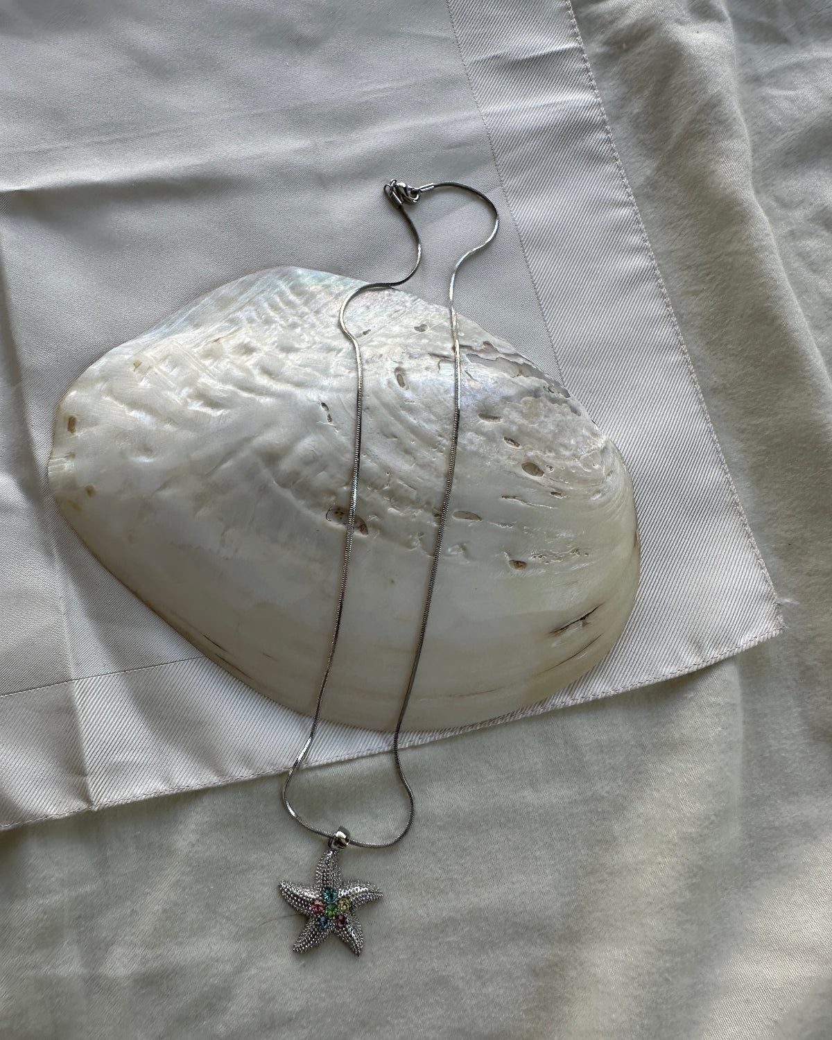 A silver-toned necklace with a starfish pendant featuring small green and pink gemstone accents, draped over a large white clam shell on a white fabric background.