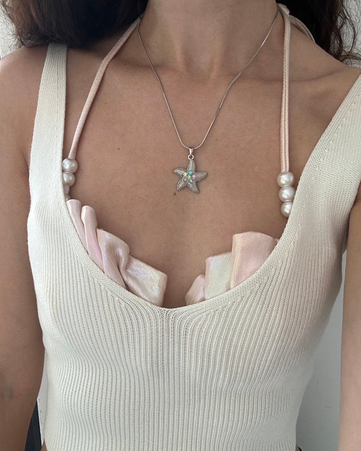 A close-up of a person wearing a silver starfish pendant necklace with a small iridescent center stone, layered over a cream ribbed camisole top with pale pink ruffled trim and pearl bead details on the straps.