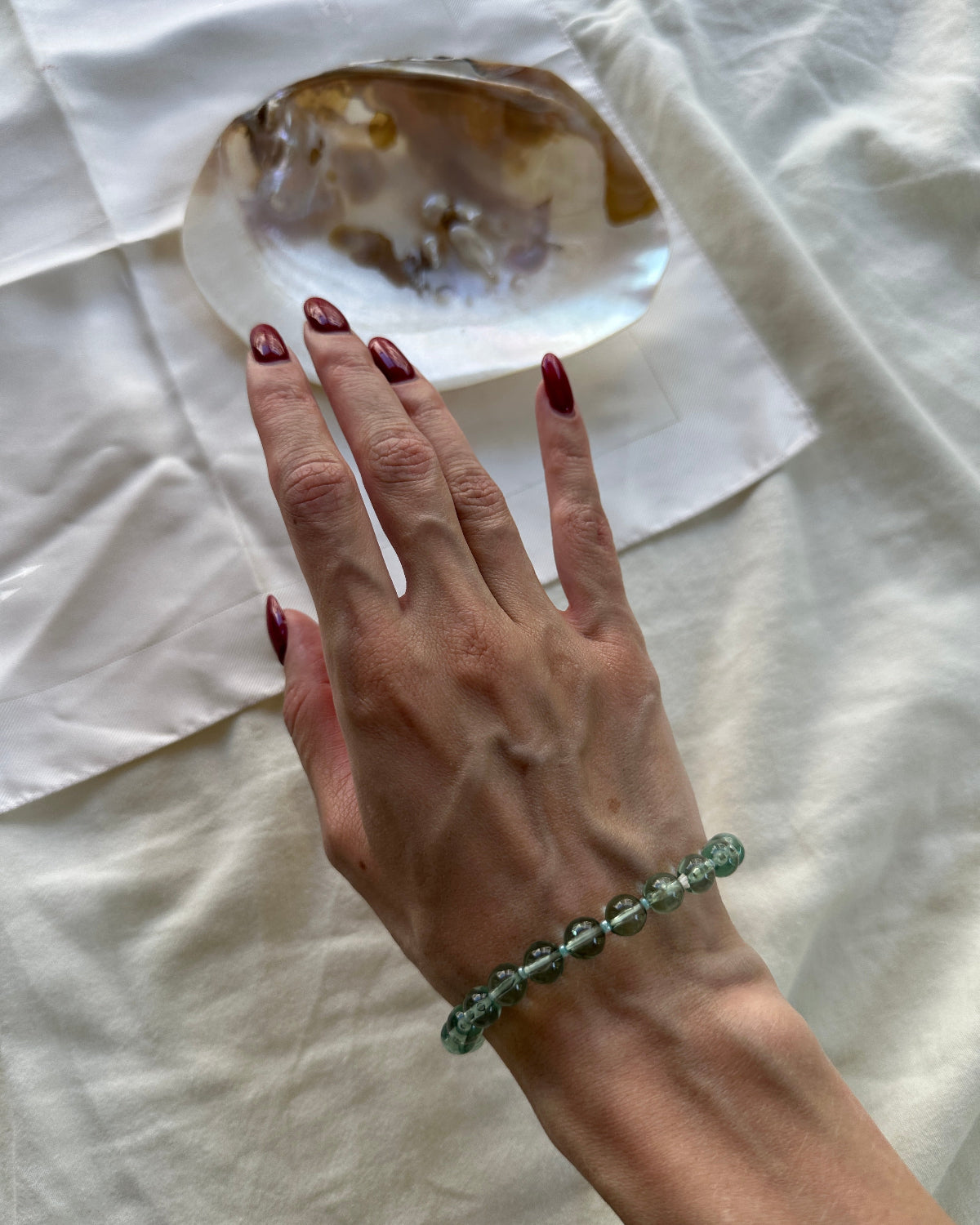 A person's hand with dark red nail polish resting on a white fabric surface next to a polished agate stone, wearing a green translucent beaded bracelet.