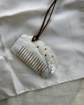 A carved white hair comb with a dark brown hanging cord, displayed on a textured off-white fabric background.
