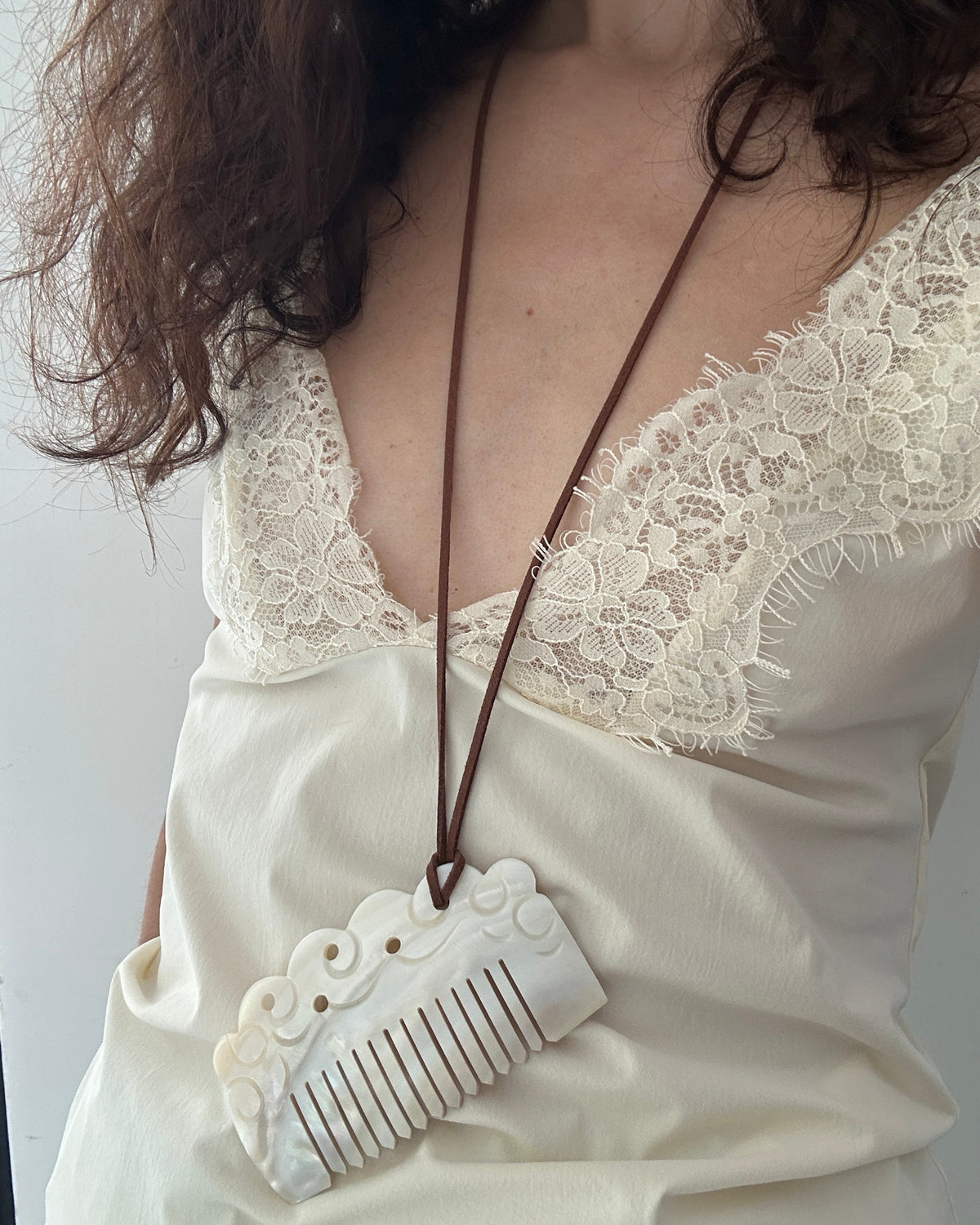 A close-up of a person wearing a cream-colored carved comb pendant necklace on a brown cord, styled over a cream lace-trim camisole.