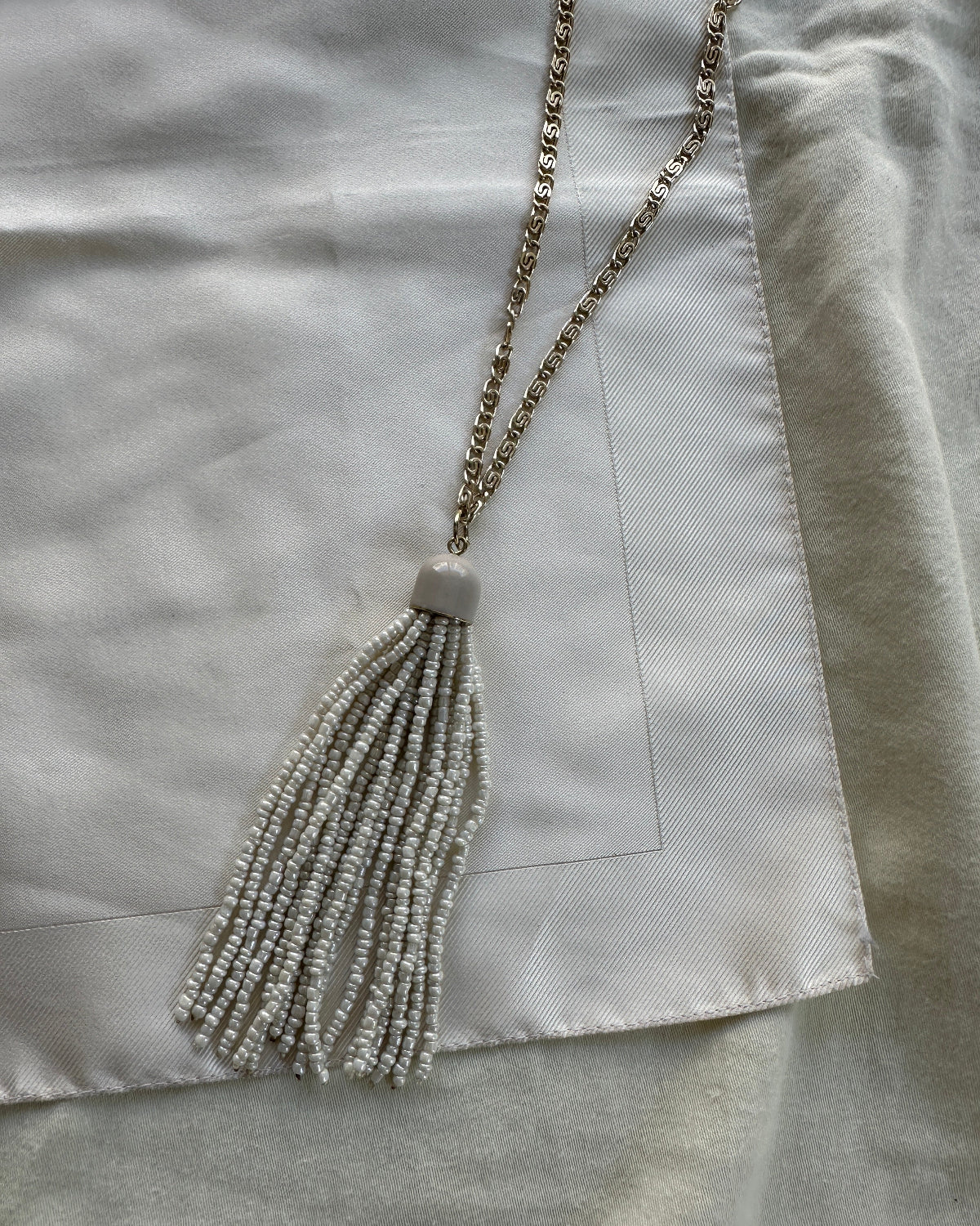 A long necklace with a delicate gold-tone chain and a large tassel made of small white beads, laid flat on a white satin fabric.