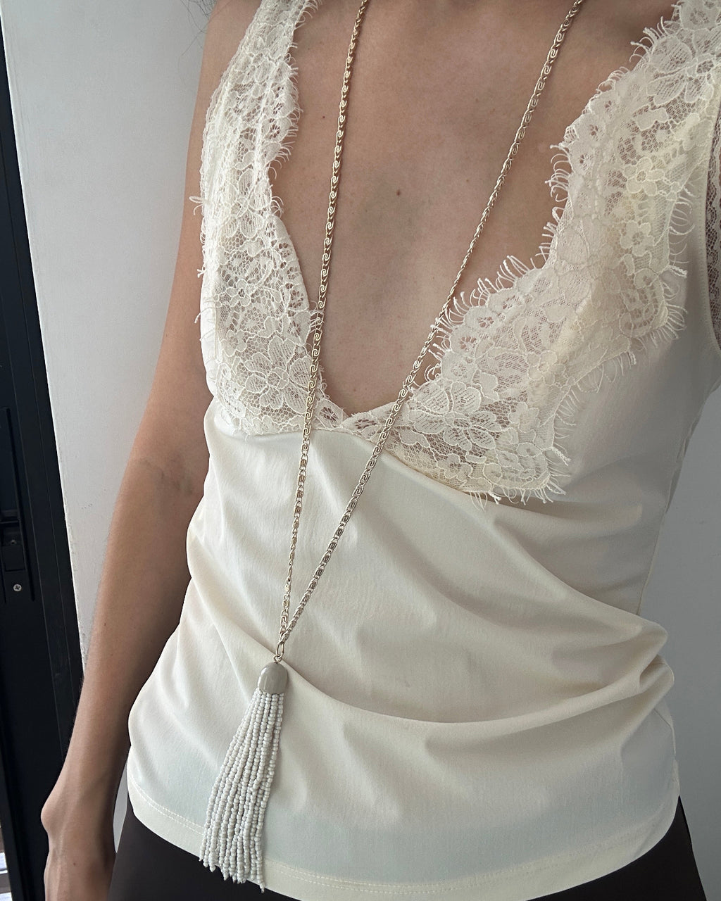 A close-up of a person wearing a cream satin camisole top with white lace trim and a long beaded tassel necklace.