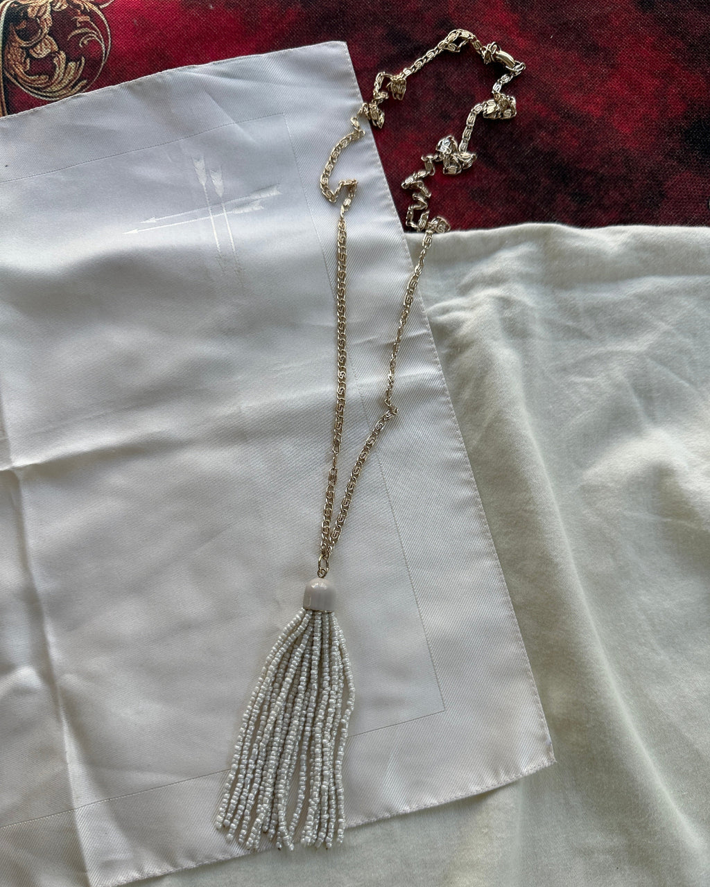 A long beaded necklace with a tassel pendant in silver and gold tones, laid out on white satin fabric with a red velvet section in the background.