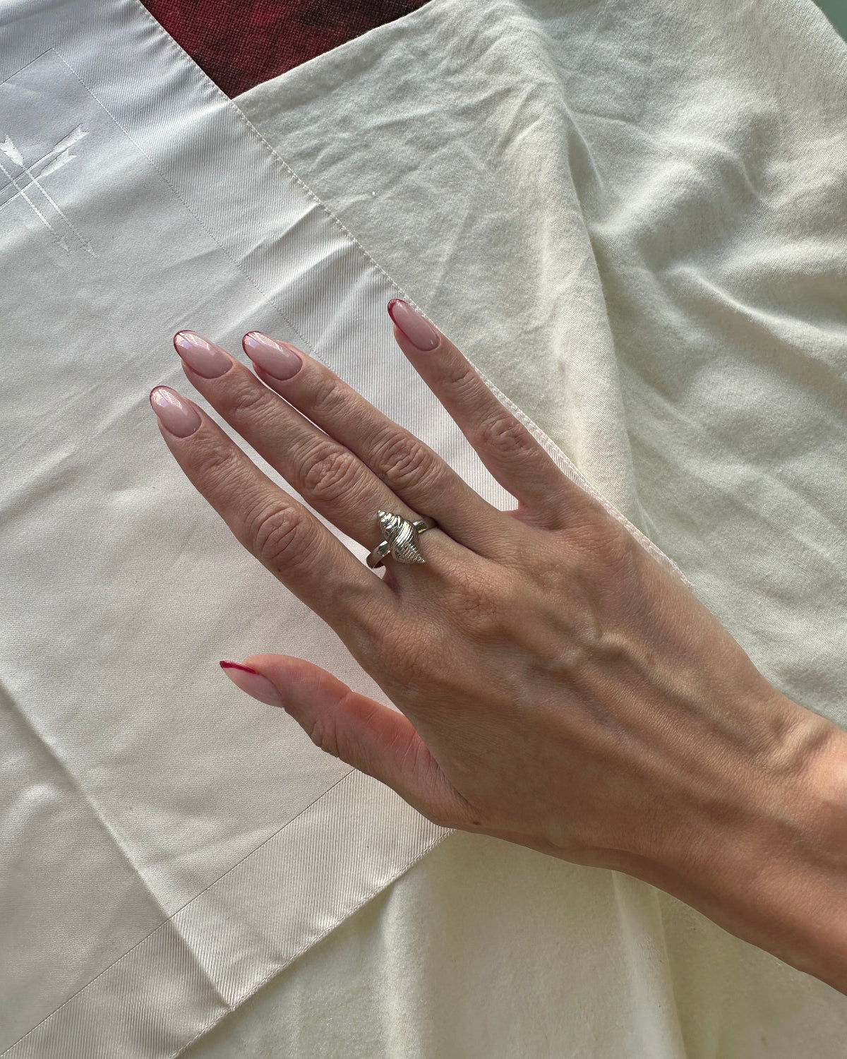 A close-up of a person's hand with manicured nails wearing a silver ring, resting on a draped cream-colored fabric.
