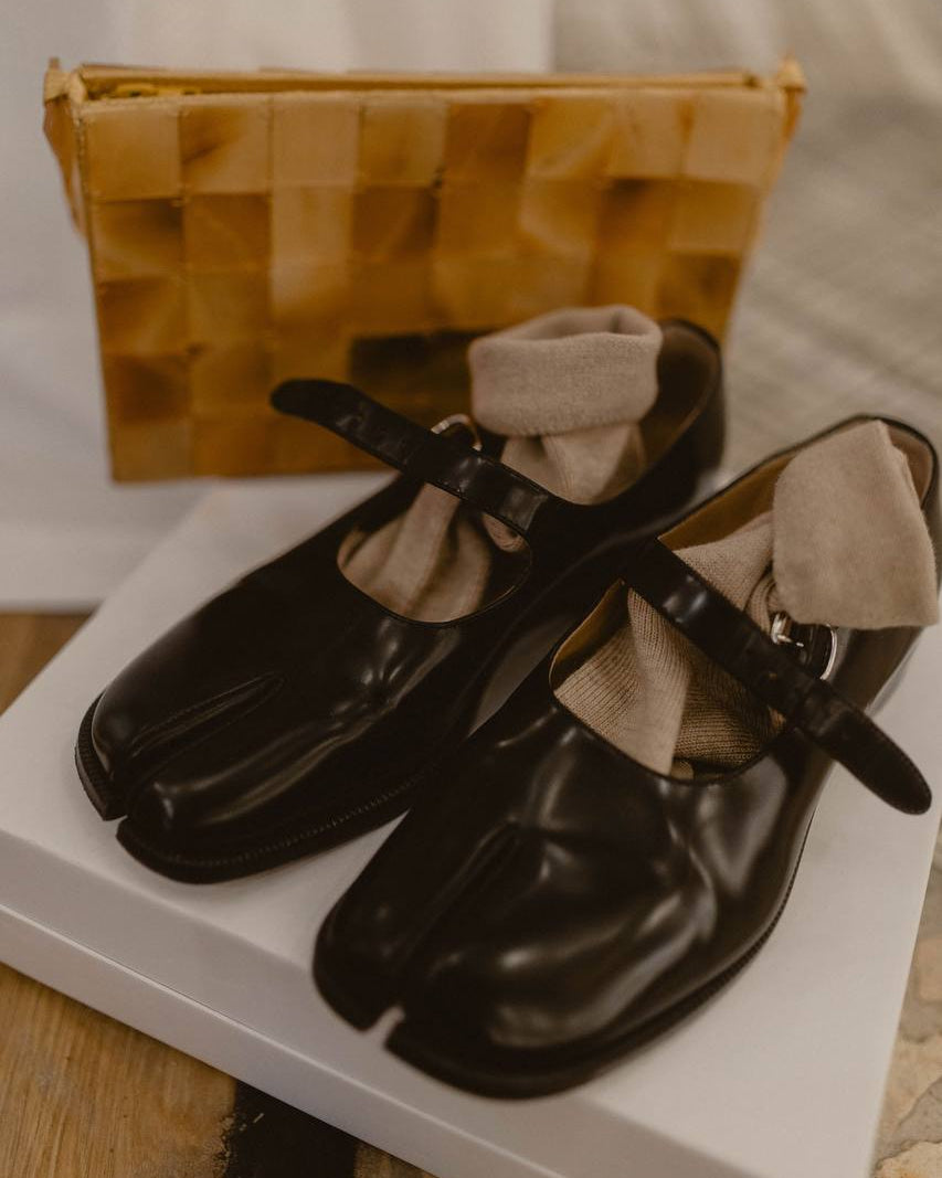 A pair of black tabi-style Mary Jane shoes with beige socks inside, displayed on a white box with a woven tan clutch bag in the background.