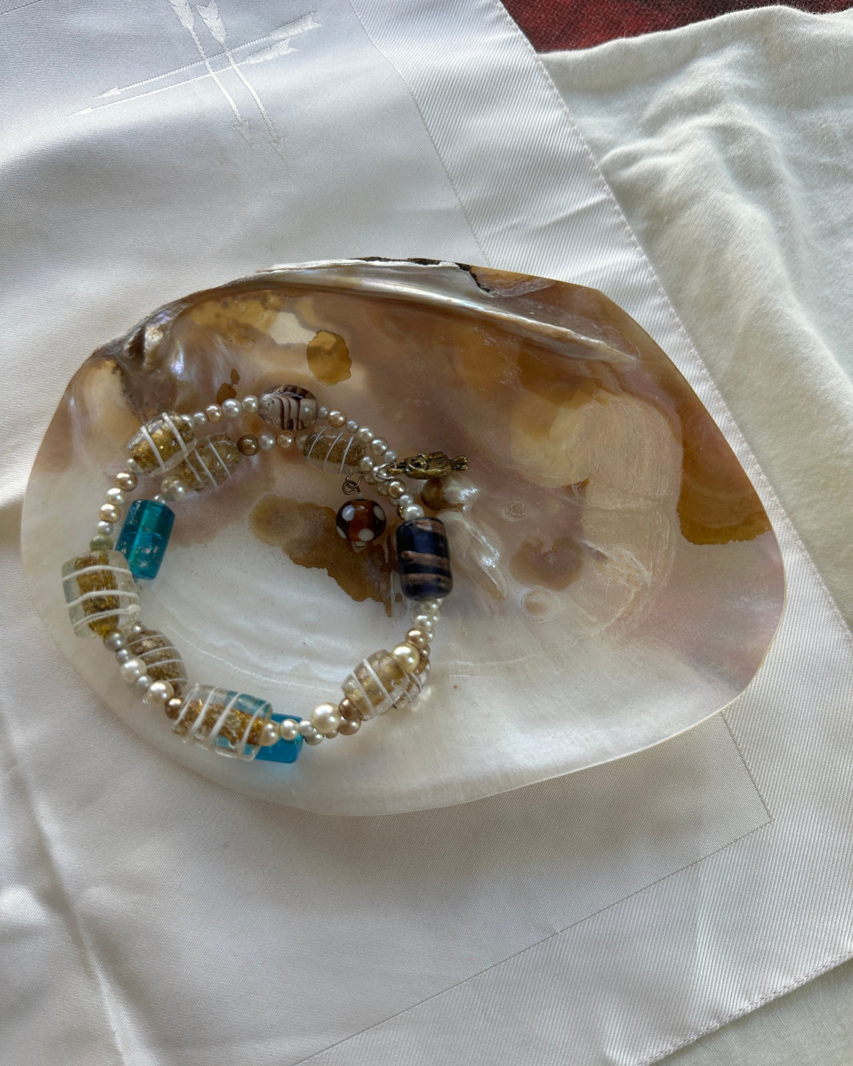 A beaded bracelet with white pearls, clear beads, and turquoise blue accents, displayed inside a polished agate shell on a white fabric background.