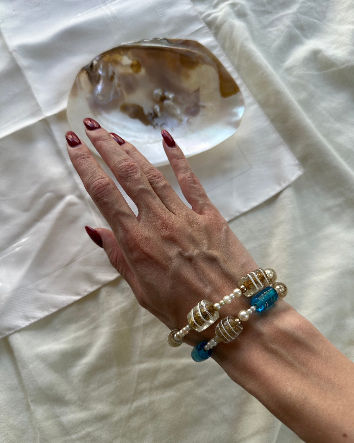 A close-up of a person's hand with dark red nail polish resting on a white fabric surface, wearing three beaded bracelets: one with white pearl-like beads, one with gold-toned textured beads, and one with blue beads.