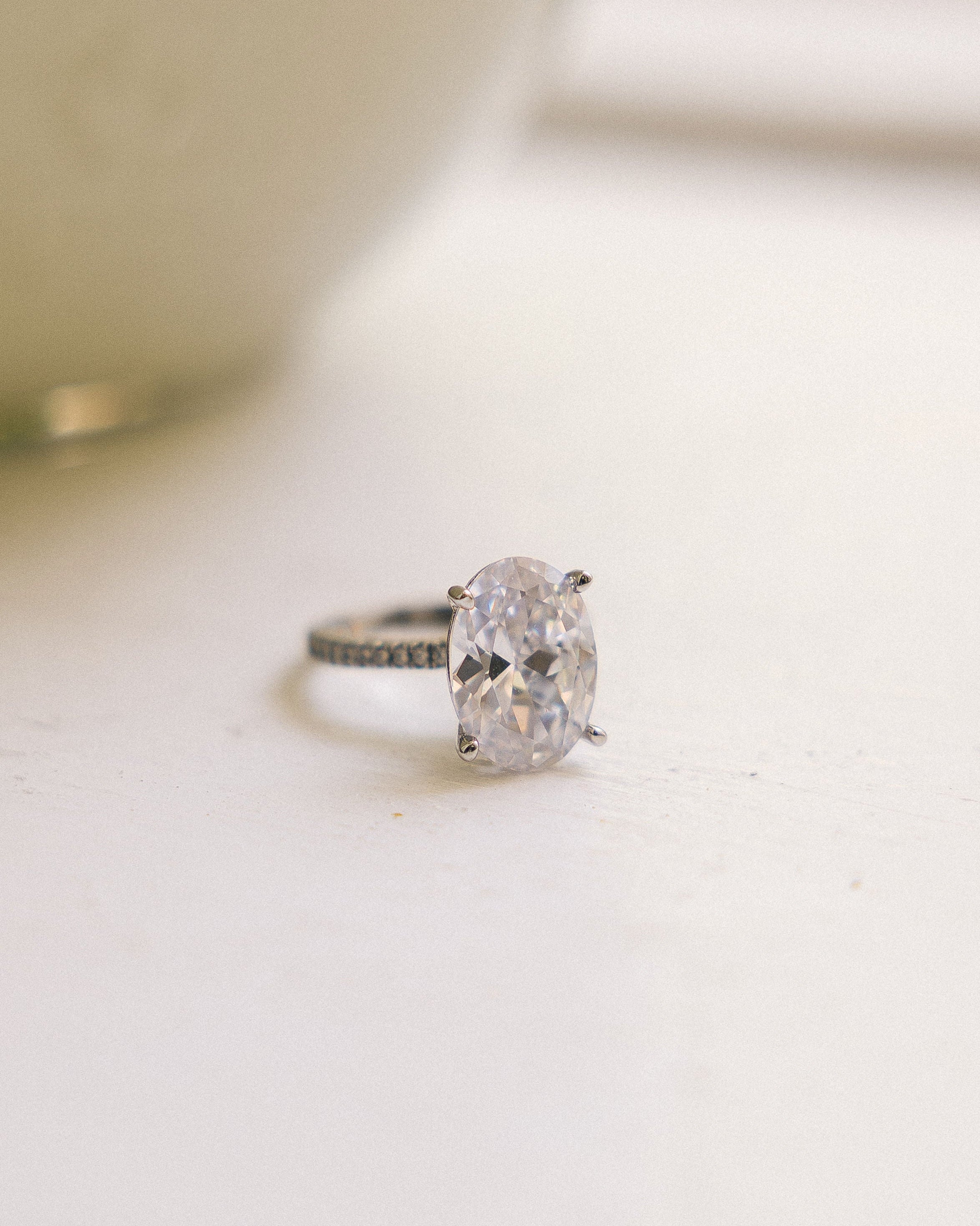 An oval-cut diamond engagement ring with a silver-toned band, resting on a light surface with a soft-focus background.