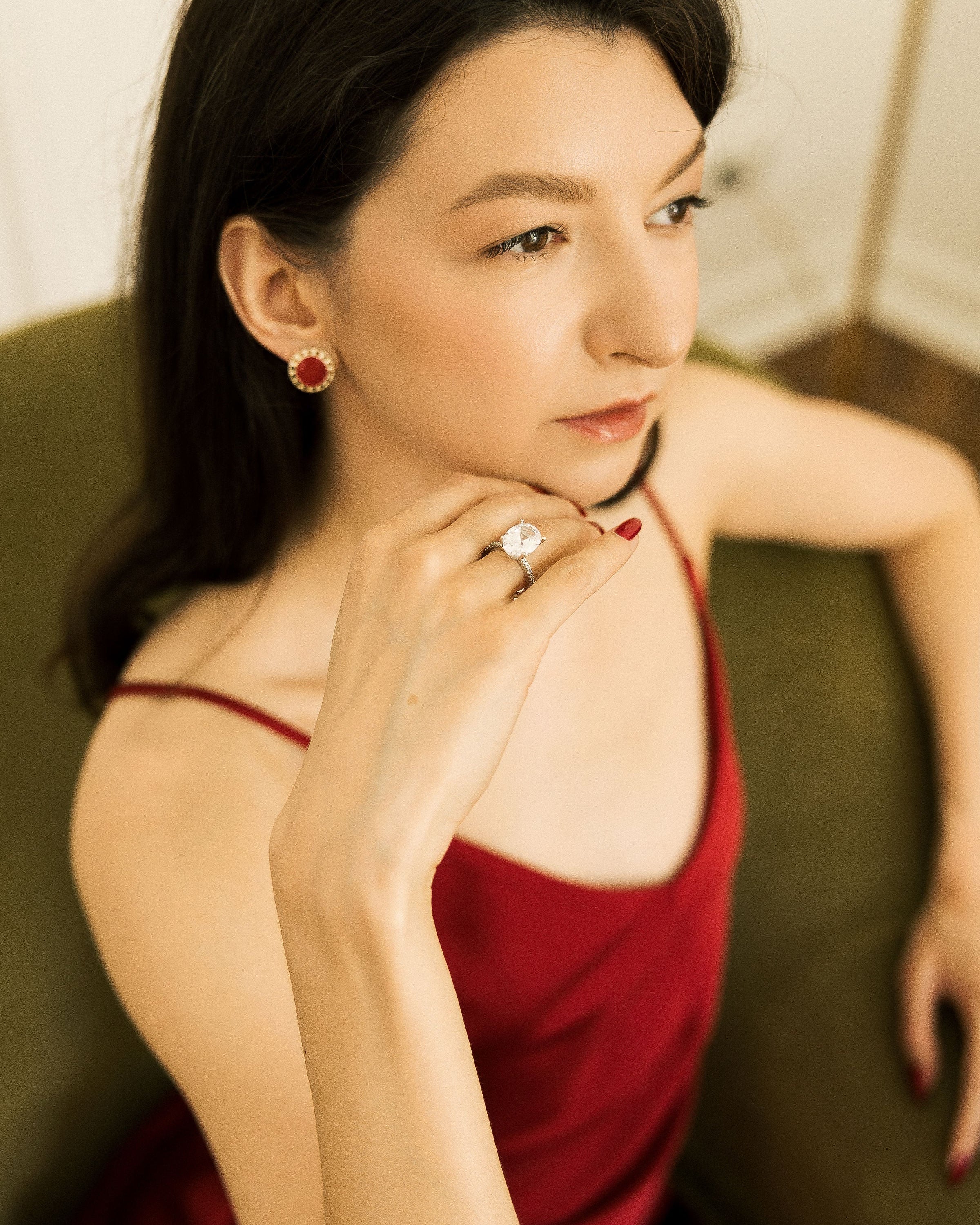 A woman with dark hair wearing a red satin camisole dress, a silver ring with a clear center stone, and matching red earrings, posing indoors against a dark olive green couch.