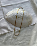 A gold chain necklace with a small white stone pendant, draped over a large white seashell on a white fabric background.