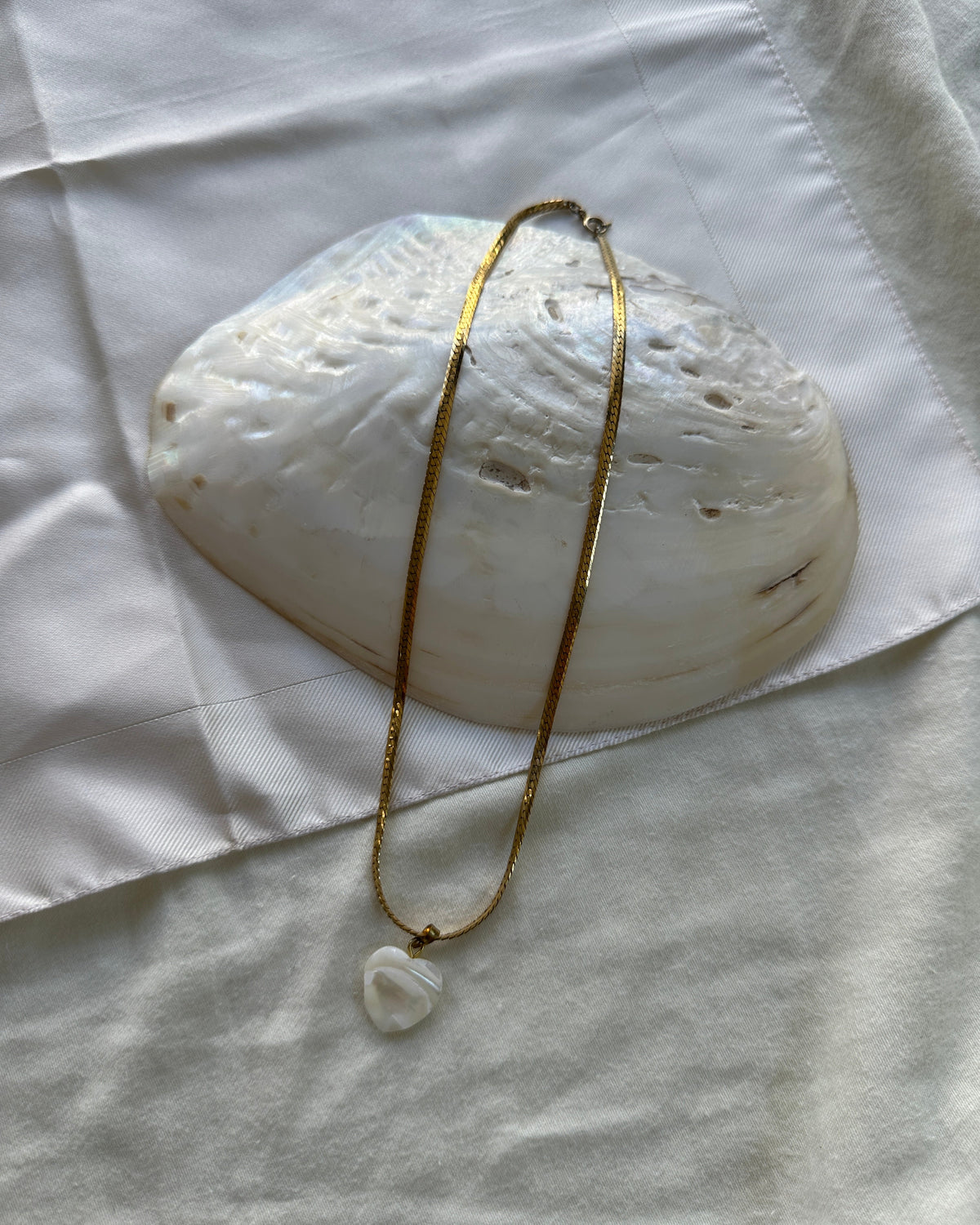 A gold chain necklace with a small white stone pendant, draped over a large white seashell on a white fabric background.