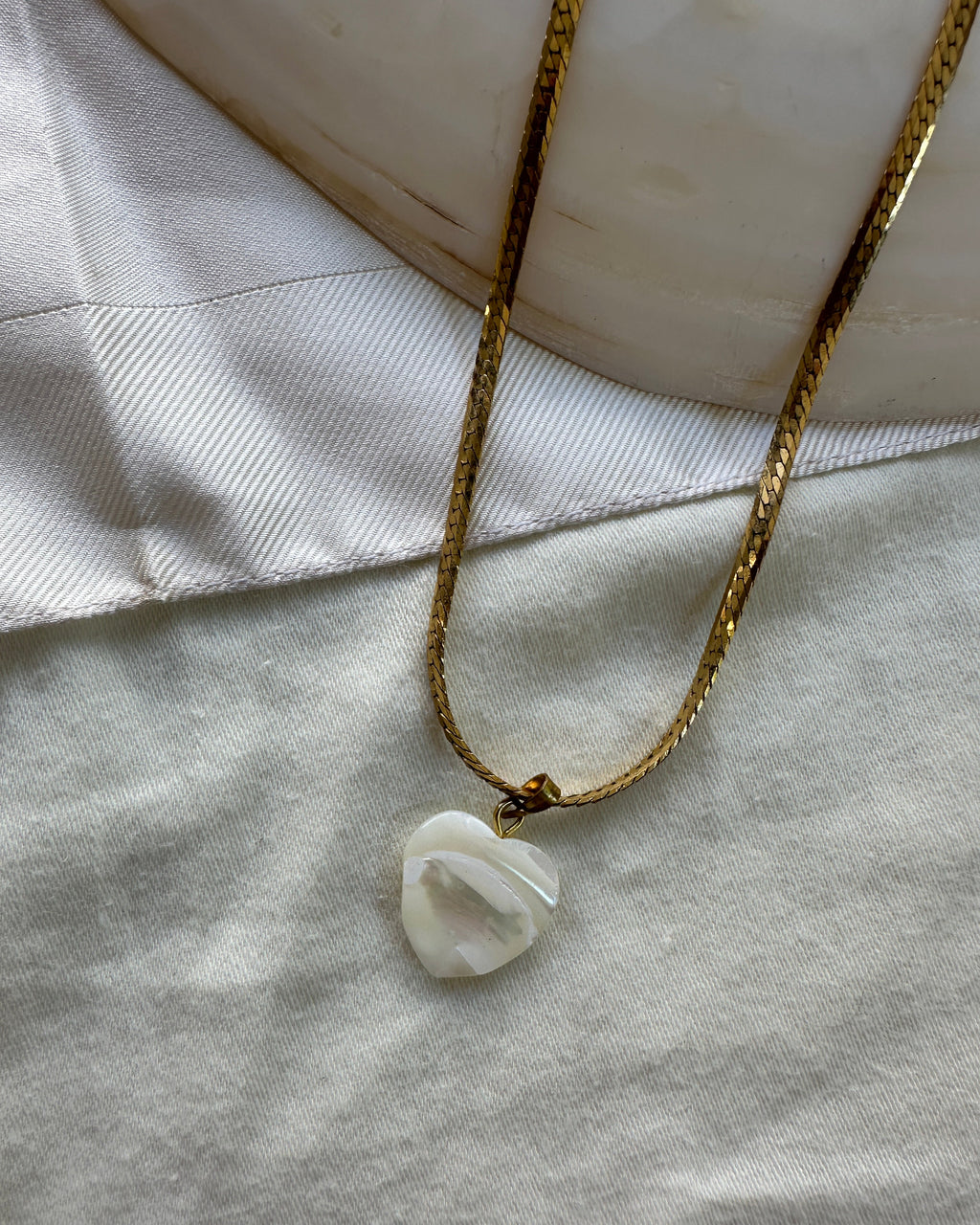 A gold chain necklace with a polished white stone pendant, styled on a light gray fabric surface next to a white satin ribbon.
