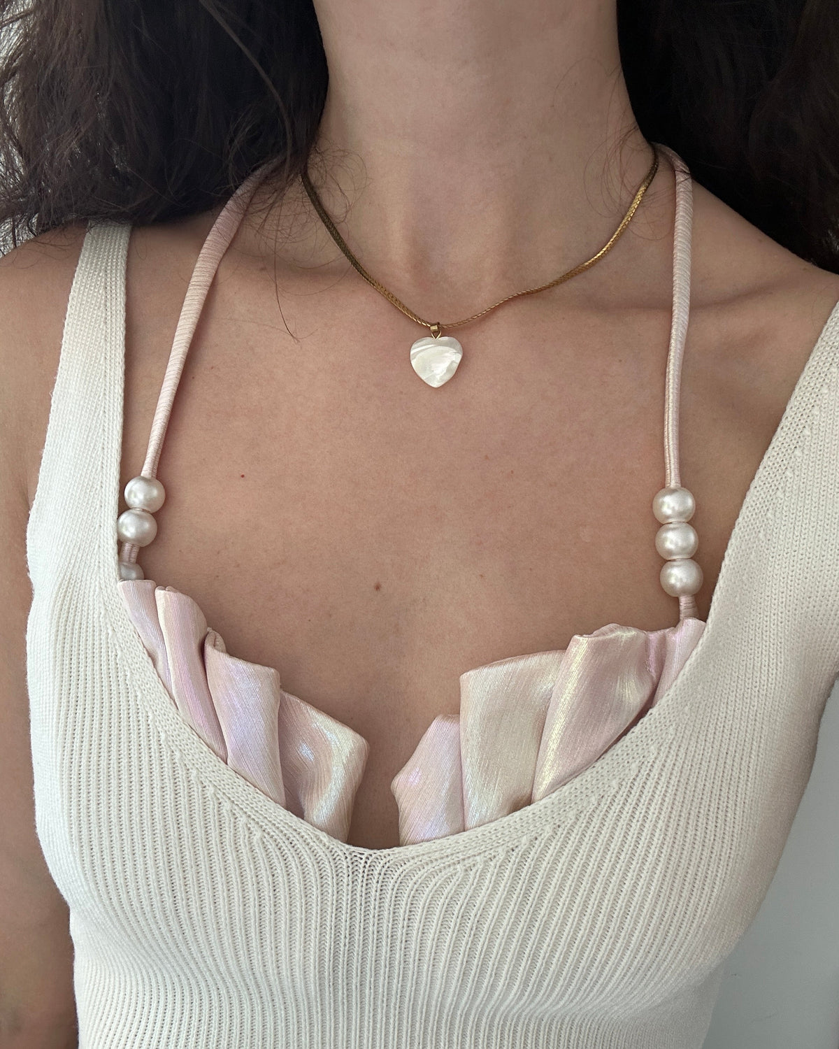 A close-up of a model wearing a delicate gold chain necklace with a small white heart pendant, layered over a gold chain. The necklace is shown on a model wearing a cream ribbed tank top with light pink satin ruffles and pearl-beaded straps.