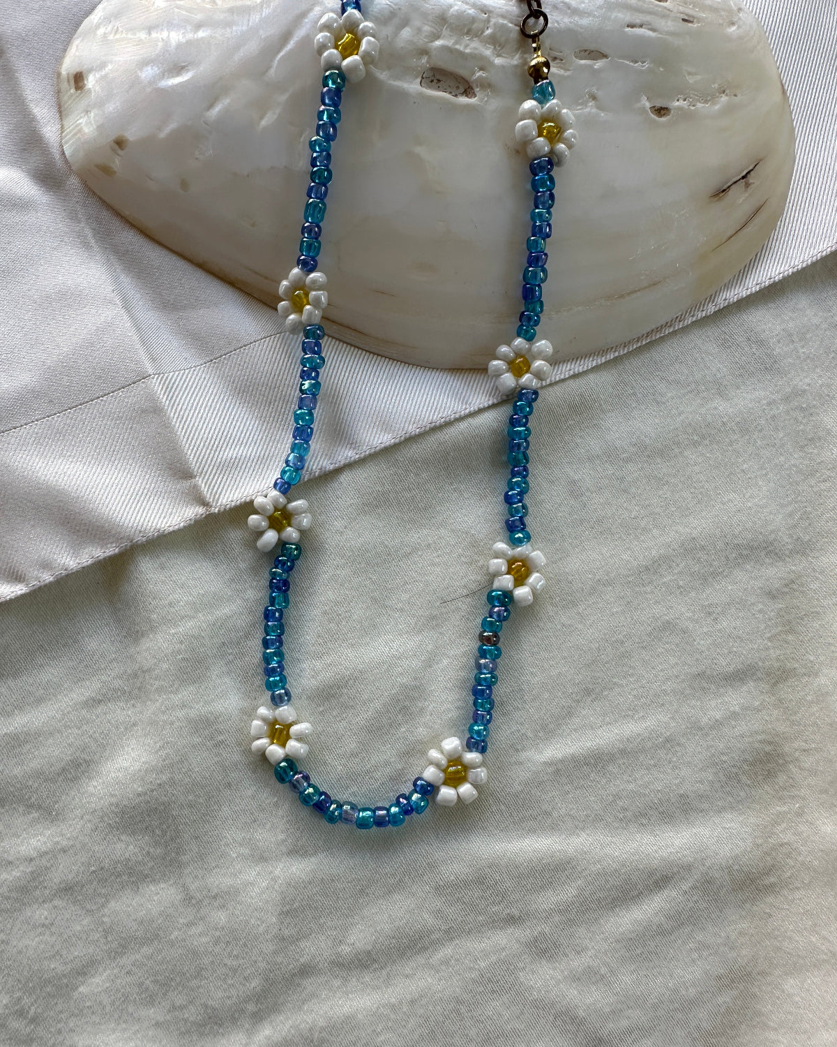 A beaded necklace featuring blue faceted beads and white daisy flowers with yellow centers, displayed on a textured white fabric surface next to a white shell.
