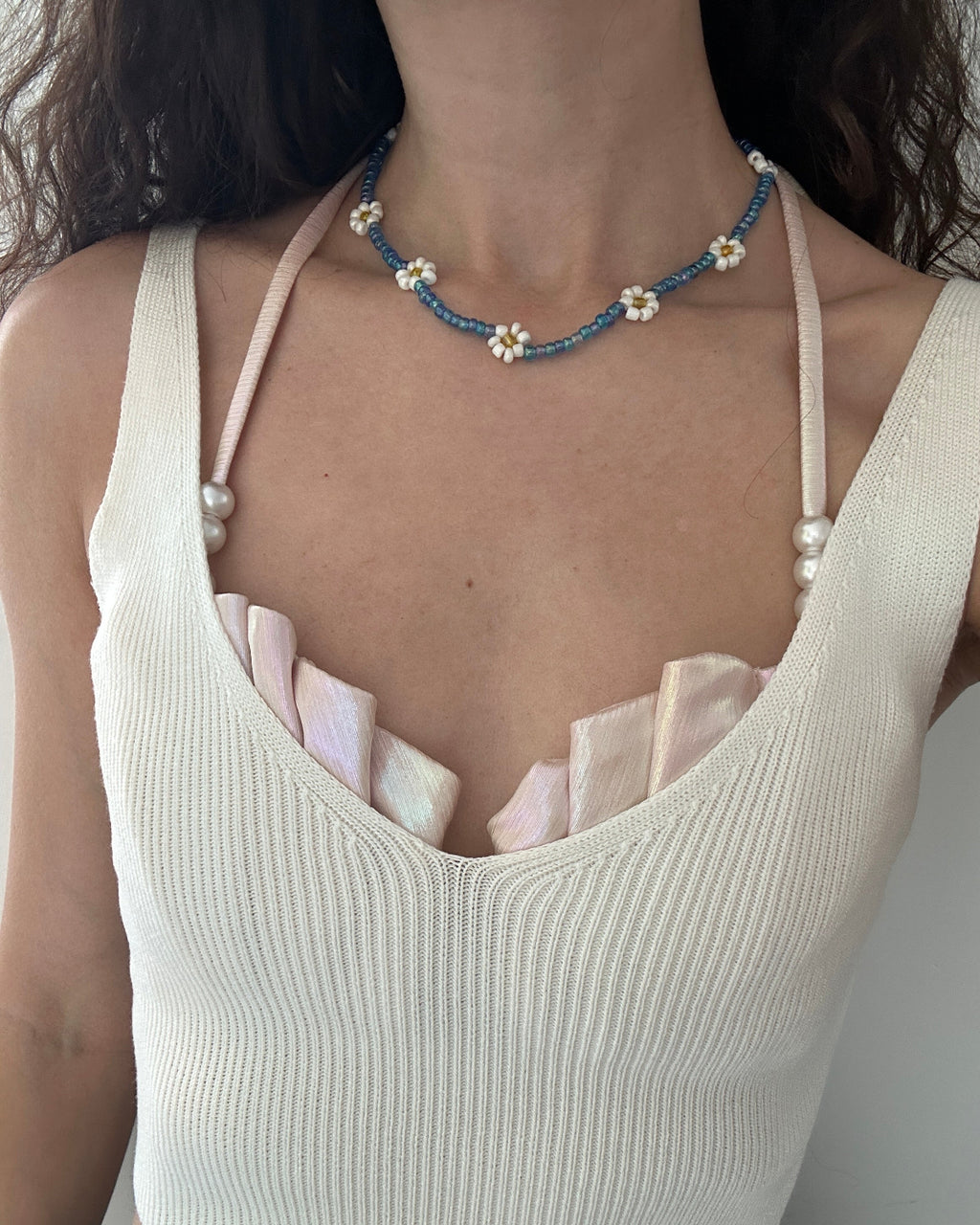 A close-up of a person wearing a delicate beaded necklace with blue beads, white pearls, and small white flower centers. The necklace is styled over a cream ribbed knit camisole top with pale pink ruffled trim.