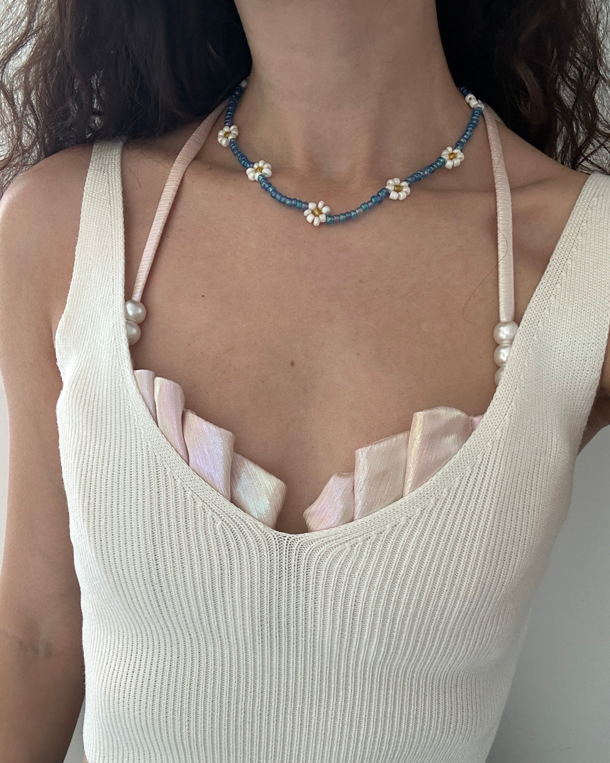 A close-up of a person wearing a delicate beaded necklace with blue beads, white pearls, and small white flower centers. The necklace is styled over a cream ribbed knit camisole top with pale pink ruffled trim.