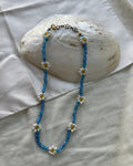 A beaded necklace featuring blue seed beads and white daisy flowers with yellow centers, draped over a large white seashell on a white fabric background.