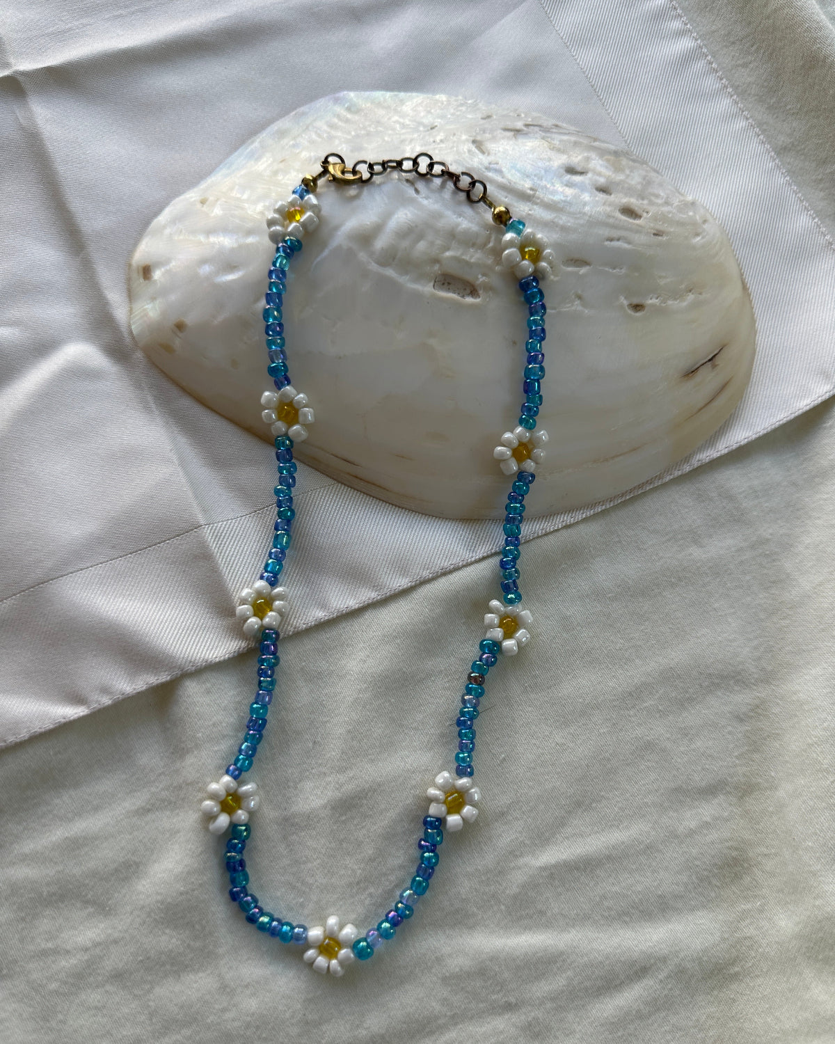 A beaded necklace featuring blue seed beads and white daisy flowers with yellow centers, draped over a large white seashell on a white fabric background.