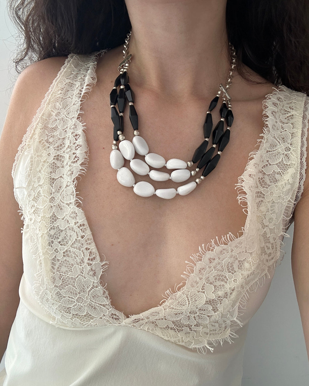 A close-up of a model wearing a multi-strand statement necklace with black faceted beads and white oval beads, styled over a cream lace-trimmed top.