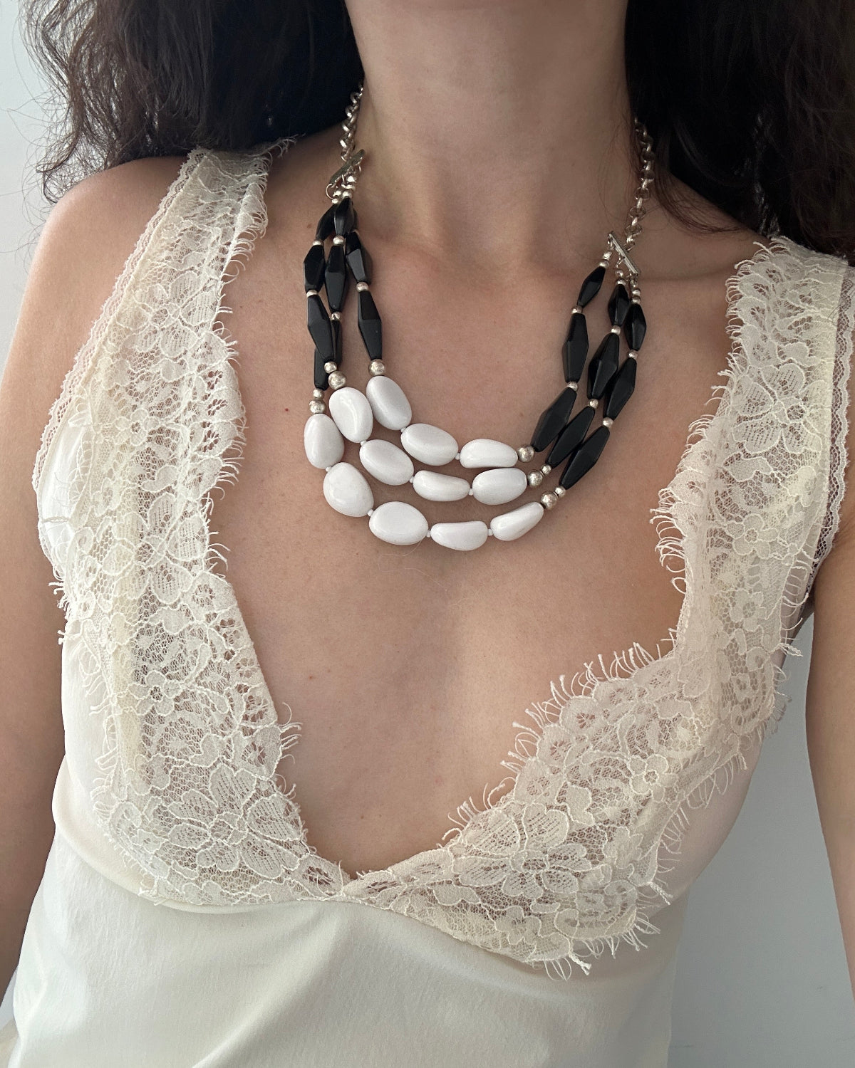 A close-up of a model wearing a multi-strand statement necklace with black faceted beads and white oval beads, styled over a cream lace-trimmed top.