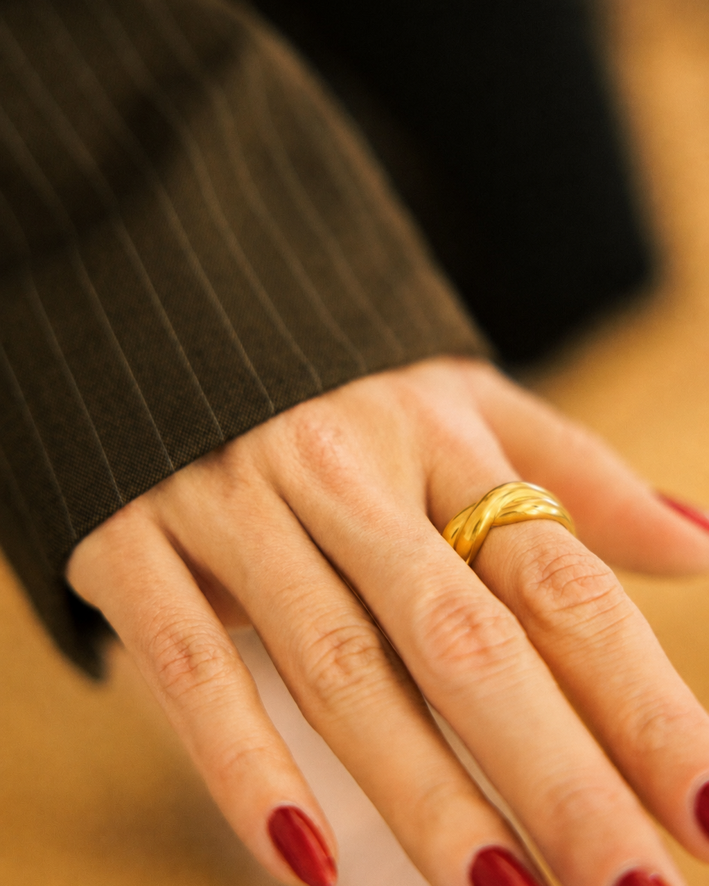 A close-up of a hand wearing a textured gold band ring, with dark red nail polish and a dark brown pinstriped sleeve visible.